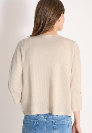 Femme portant un pull côtelé beige clair à manches trois-quarts et un jean bleu, vue de dos sur un fond uni.