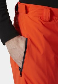 Oranje waterdichte broek met een zijzak met rits, riemlussen en een gladde textuur. Close-up geeft een deel van een zwart langemouwen shirt weer.