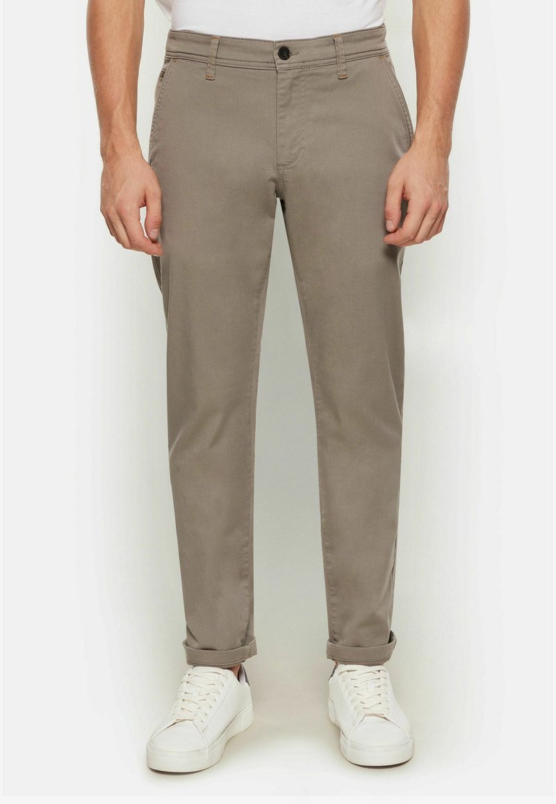Khakifarbene Chinos mit schmaler Passform, ausgestattet mit einem Knopfverschluss, Fronttaschen und aufgerollten Säumen, kombiniert mit weißen Sneakers auf einem schlichten Hintergrund.