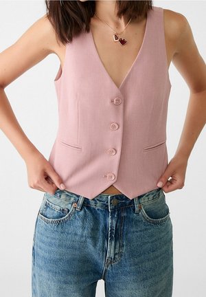 Woman wearing a sleeveless dusty pink buttoned vest and blue denim jeans, adjusting the hem of the vest with both hands.