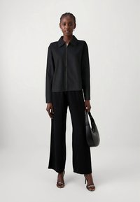 Black zip-up jacket with a collar, paired with wide-leg black trousers. Model holds a black handbag and wears silver strappy heels.