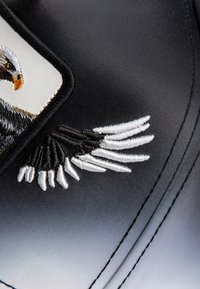 Tela negra con un parche de águila con bordado detallado; cabeza y ala del águila en tonos negros, blancos y dorados con textura suave.