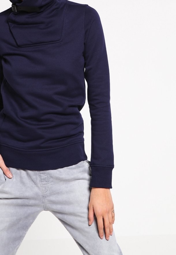 Person wearing a dark blue long-sleeve top with a high neck and light gray pants, posing against a white background.