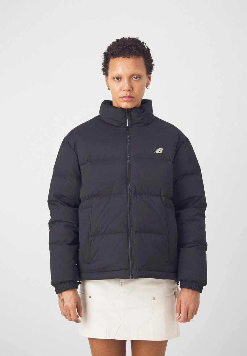 Person in einer schwarzen Daunenjacke mit einem Frontreißverschluss und Taschen, kombiniert mit einem weißen Rock, vor einem schlichten Hintergrund stehend.