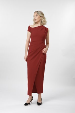 Γυναίκα με ασύμμετρο, σκουροκόκκινο, αποκαλυπτικό φόρεμα με ανοίγματα στο πλάι, φοράει μαύρα τακούνια, στέκεται μπροστά σε απλό ανοιχτό φόντο.