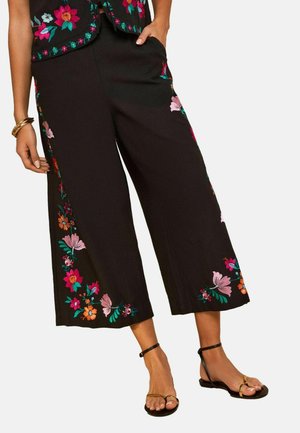 Pantalon court noir à jambes larges avec broderie florale colorée le long des côtés et des ourlets, porté avec des sandales dorées et des bracelets dorés.