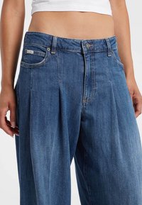 Pantaloni in denim blu con un design plissettato, stile a cinque tasche e chiusura con bottone. Il tessuto ha una finitura liscia e leggermente testurizzata.