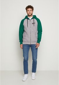 Grauer Reißverschluss-Hoodie mit grünen Ärmeln, ausgestattet mit Kordeln und seitlichen Taschen. Kombiniert mit blauen Jeans und weißen Sneakers.