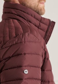 Mężczyzna w bordo pikowanej kurtce puchowej z wysokim kołnierzem i małym okrągłym logo "Superdry" na górnej części ramienia.