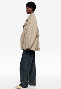 Naine seisab profileerimises, seljas suur beezist villane tikandiga jakk, laiad tumedates pesu valuudest teksad ja kreemvalged terava ninaga kingad.