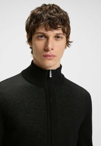 Svart zip-up-tröja med hög krage, med strukturerad stickad material och en slät dragkedja. Tajt passform, minimalistiska mönster.