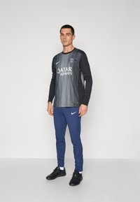 Pánska dlhá športová dres v čiernej farbe s vertikálnymi sivými pruhmi a logom PSG, doplnený o rebrované nohavice v námorníckej modrej farbe a čierne športové topánky.