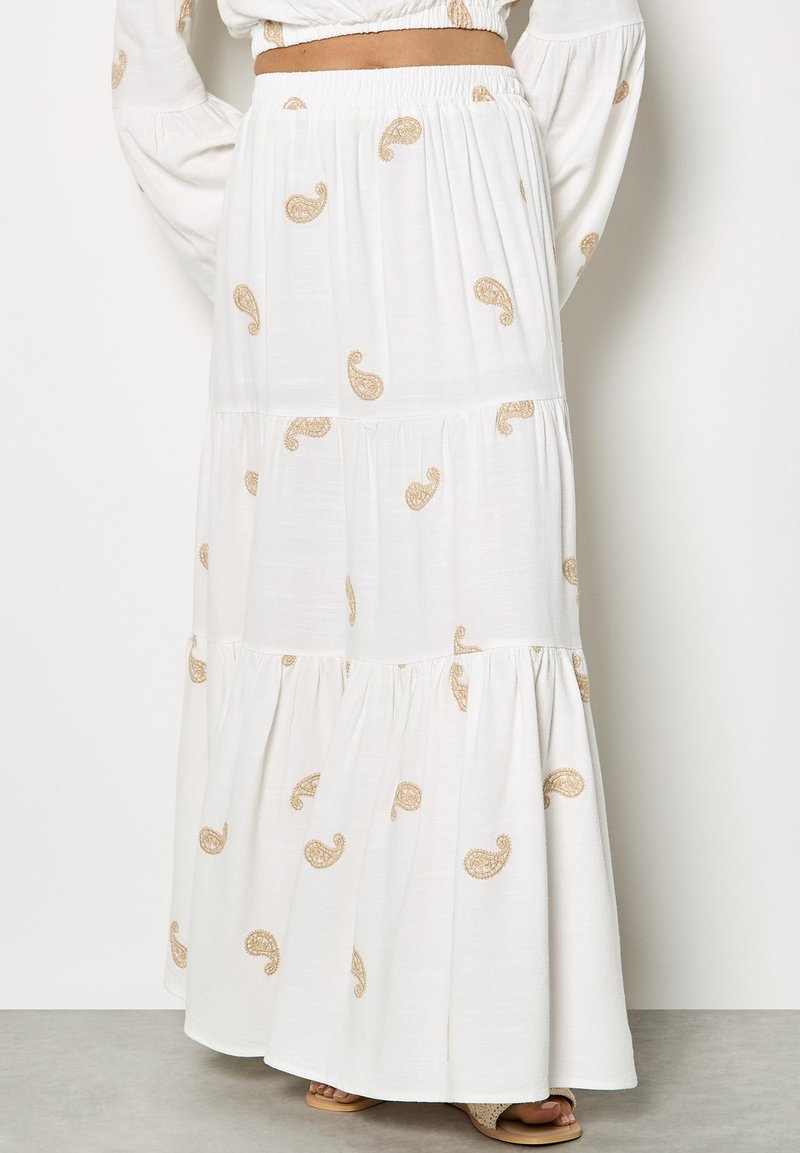 Jupe longue blanche à étages avec taille élastique, ornée de motifs cachemire marron clair, portée avec un haut blanc assorti et des sandales.