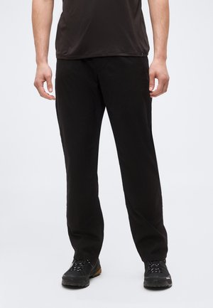 Férfi, fekete sportnadrágban és fekete túracipőben, világos háttér előtt áll.