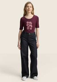Maroon fitted top with short sleeves and abstract pink line design, paired with blue wide-leg jeans and white canvas shoes.