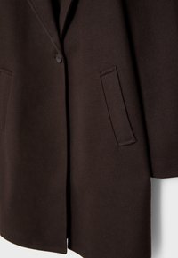 Manteau marron foncé avec une fermeture à un seul bouton et une poche fendue verticale sur le côté droit devant, en tissu texturé.