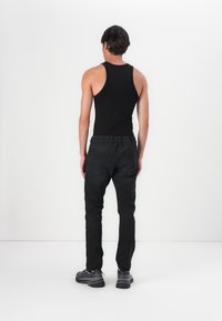 Črn top brez rokavov v kombinaciji s črnimi hlačami, s telesno prileganjem in elastičnim pasom. Model nosi sivo obuvalo, prikazan s hrbta.