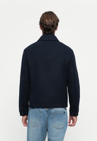 Man with brown hair wearing a navy jacket and light blue jeans, shown from the back against a plain white background.