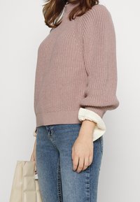 Pull tricoté rose clair avec un col montant et des manches ballon, associé à un poignet de chemise blanc à volants, porté avec un jean bleu.