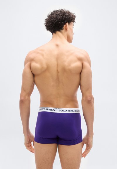 Μυώδης άνδρας, ο οποίος φαίνεται από πίσω, φοράει μοβ μποξεράκια Polo Ralph Lauren, σε απλό λευκό φόντο.