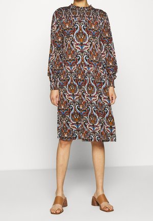 Kvinde iført knælængde, langærmet kjole med flerfarvet paisleymønster og høj smocket halsudskæring, kombineret med tan åbne blockhælsandaler.