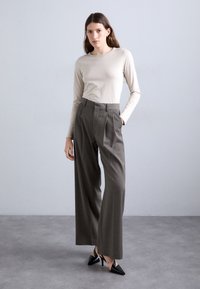 Haut long beige clair associé à un pantalon tailleur gris foncé à taille haute et plis, avec des escarpins noirs à bout pointu. Tissu lisse et coupe ajustée.