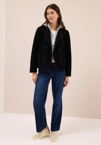 Kvinna stående i blå jeans, vita sneakers, vit topp under en grå zip hoodie och en svart dubbelknäppt jacka i en neutral studio.