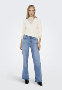 Light cream sweater with lace v-neck, paired with light blue flared jeans. Model wears black ankle boots. Smooth texture; casual design.