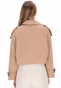 Kort beige trenchcoat med stor krage, dekorativa knappar på ärmarna och två sidobeltesdetaljer. Mjukt tyg med slät textur.