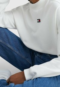 Sudadera blanca con cuello alto, que presenta un parche rectangular con el logo en rojo, blanco y azul. Combinada con vaqueros de mezclilla azules.