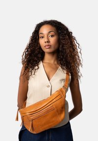 Tan läder crossbody-väska med dragkedjestängning och fransdetaljer, med en rundad form och justerbar rem. Modellen bär en beige topp.