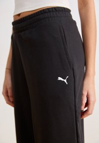 Črne bombažne trenirke z elastičnim pasom, ki vključujejo stranske žepa in bel akcent s Puma logotipom na levi nogi.