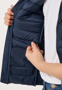 Une personne portant une veste matelassée bleu marine et une chemise blanche ouvre la poche intérieure, montrant la doublure et les détails de fabrication.