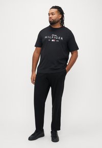 Μαύρο μπλουζάκι με λευκό λογότυπο "Tommy HILFIGER", σε συνδυασμό με μαύρο παντελόνι και μαύρα αθλητικά παπούτσια. Απλή, casual εμφάνιση σε σκούρες αποχρώσεις.