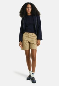 Blazer navy con costuras blancas, suéter de punto de cable navy, pantalones cortos caquis, mocasines navy y calcetines de tobillo a rayas. Composición de atuendo casual.