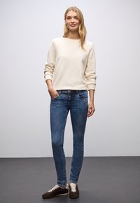 Sudadera crema con puños de canalé, jeans ajustados azules con detalles en los bolsillos, y zapatillas de ante marrones con cordones crema. Conjunto sencillo y casual.