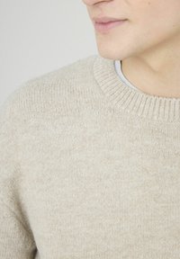 Pull beige à maille texturée, avec un col rond côtelé et un tissu doux, partiellement visible sur un fond uni.
