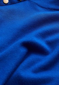 Blå stickad tröja med en mjuk yta, ribbad krage och två ljusa knappar vid halsen.