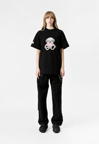 Camiseta negra de gran tamaño con un gráfico de oso texturizado en rosa y gris. Combinada con pantalones cargo negros y calzado texturizado negro.