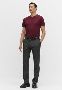 Mand står iført en burgunder kortærmet skjorte, grå skræddersyede bukser med et sort bælte og sorte sko mod en ensfarvet baggrund.