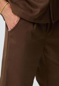 Pantalon marron texturé avec une poche latérale et une coupe ample. Le tissu semble lisse avec un léger éclat, adapté pour une tenue décontractée ou formelle.