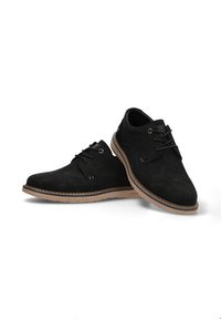 Zapatos de ante negro con punta redonda, que cuentan con un diseño de cordones, suela en contraste en color marrón claro y sutiles detalles de costura en la parte superior.