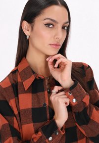 Camicia a maniche lunghe a quadri in arancione e nero, con chiusura a bottoni sul davanti, colletto e dettagli in metallo alle maniche.