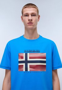 Modrá krátká tričko s krátkým rukávem, na kterém je grafika norské vlajky, s textem "NAPAPIJRI" nad designem a "geographic" pod ním.