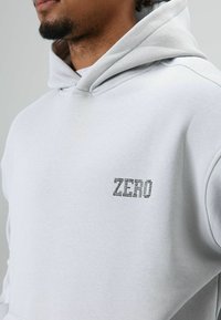 Lichtgrijze hoodie met een geribbelde kraag en een zwarte "ZERO"-logo met strass steentjes op de linkerborst. Zachte, geborstelde textuur.