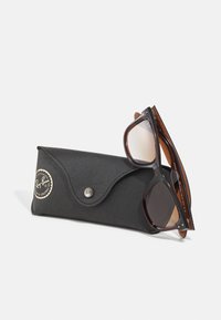 Ochelari de soare cu nuanțe de brown într-un capac texturat negru. Capacul are un sistem de închidere prin clips și un logo circular auriu. Ochelarii au un design elegant și lentile colorate.