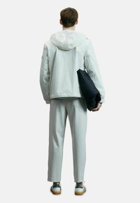 Veste légère gris clair avec capuche, associée à un pantalon assorti, tenant une doudoune noire. Des chaussures décontractées complètent le look.