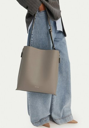 Persona vistiendo una blazer de rayas príncipe de Gales y jeans de pierna ancha, sosteniendo un gran bolso tote de cuero gris con correa para el hombro.