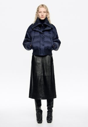 Chaqueta acolchada azul marino con cuello alto y dobladillo acanalado, combinada con una falda larga de cuero negra. Texturas suaves y detalles mínimos de herrajes.