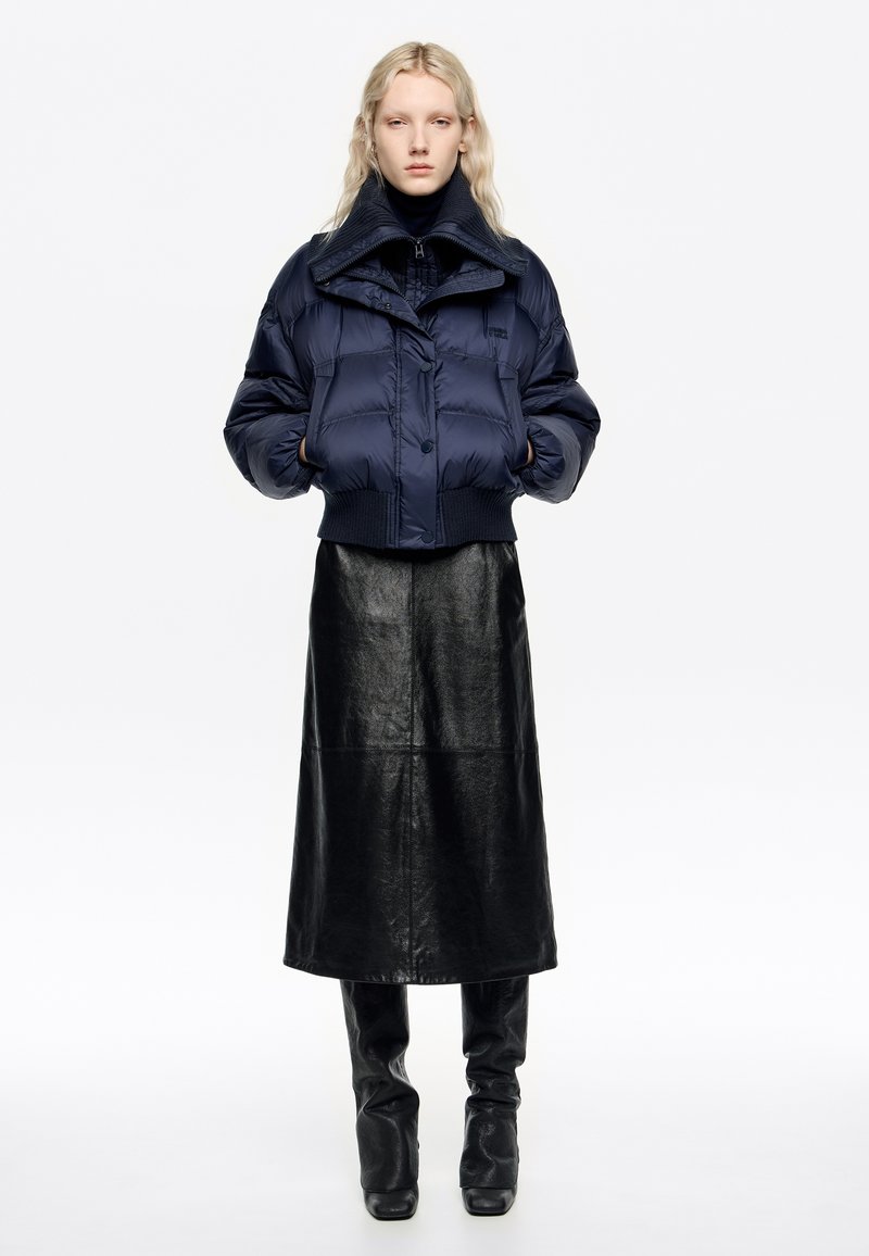 Granatowa kurtka puchowa z wysokim kołnierzem i prążkowanym dołem, zestawiona z czarną, skórzaną spódnicą maxi. Gładkie tekstury i minimalne detale z metalowymi elementami.